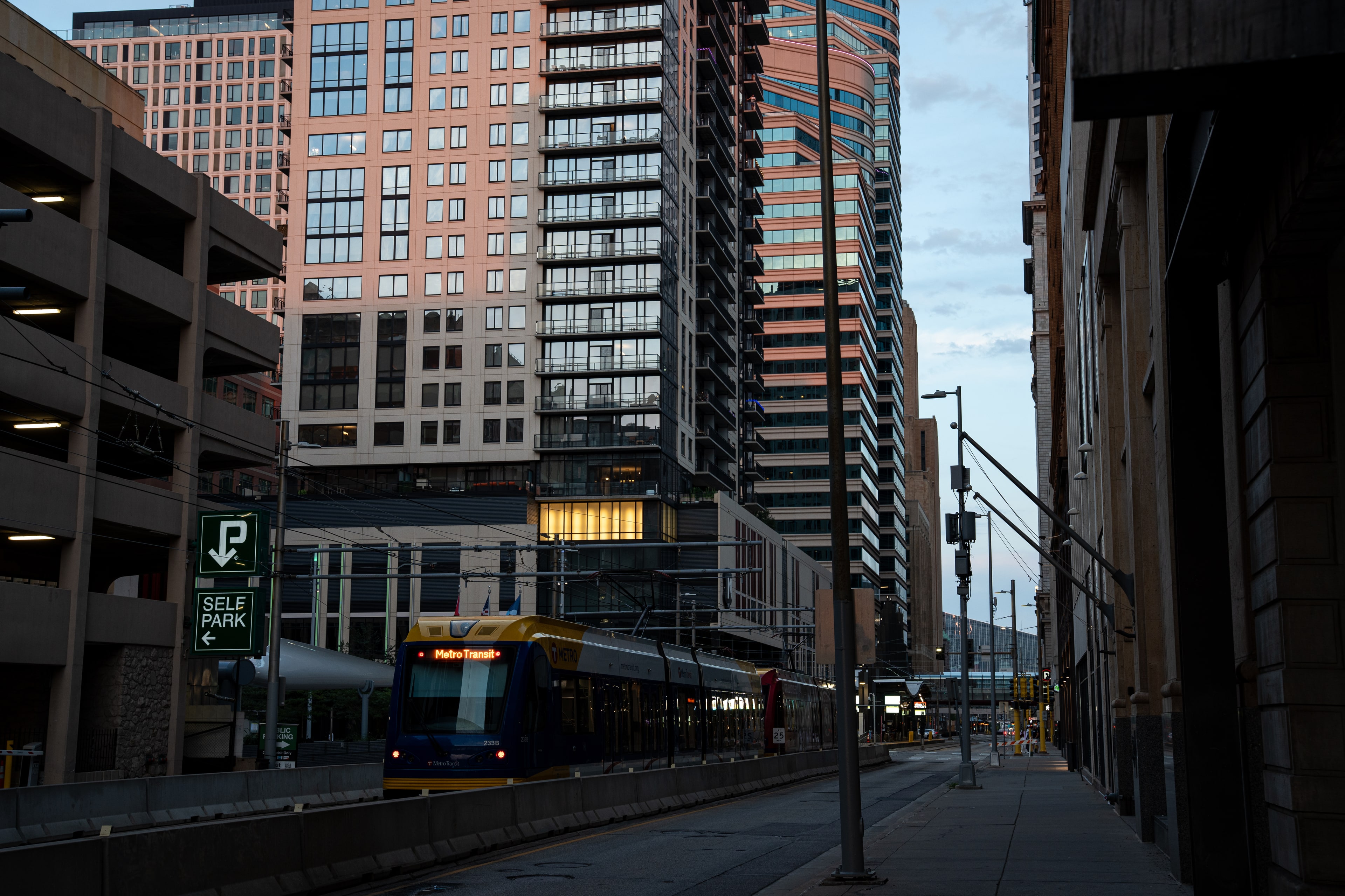 A bus pulling away beneath residential towers at dusk, the street falling into shadow.
