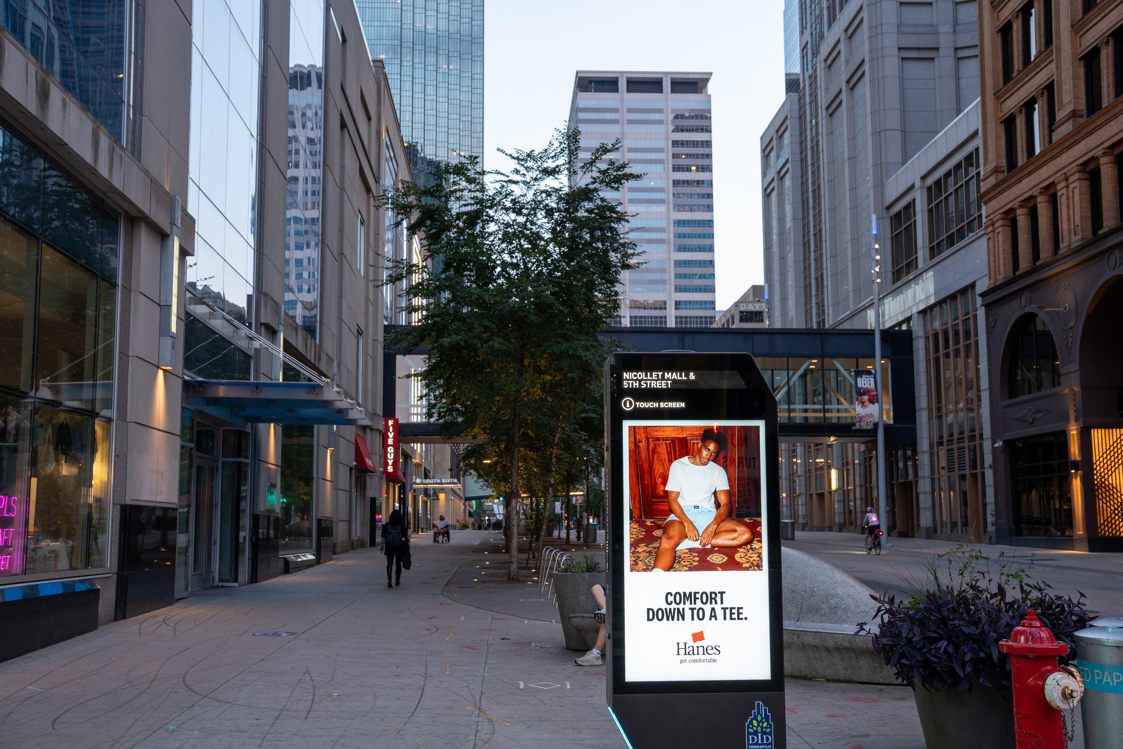 A lit digital kiosk standing on a tree-lined pedestrian mall as the surrounding towers darken.