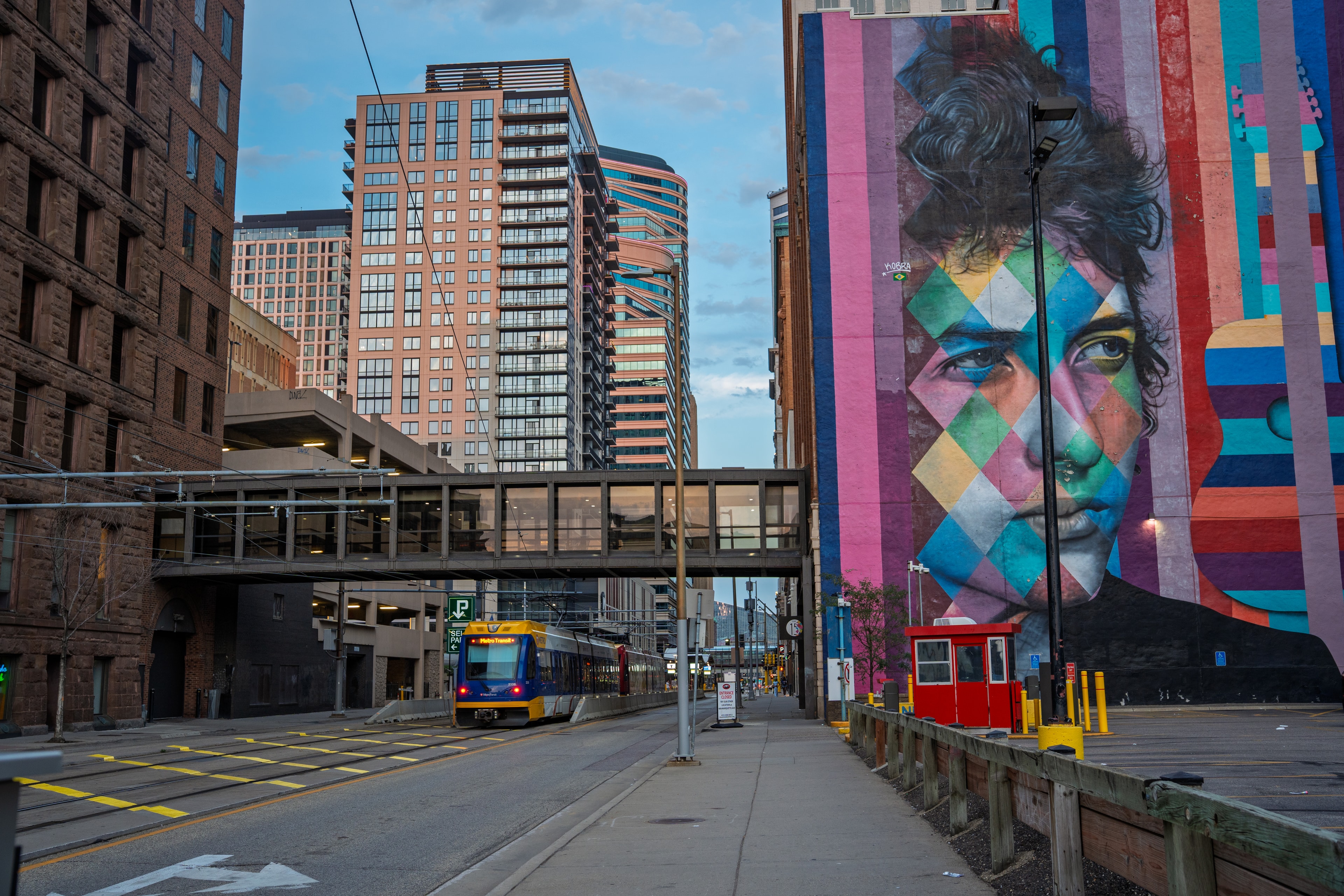 A large painted mural of Bob Dylan covering the side of a brick building, with a light-rail train and downtown towers beyond.