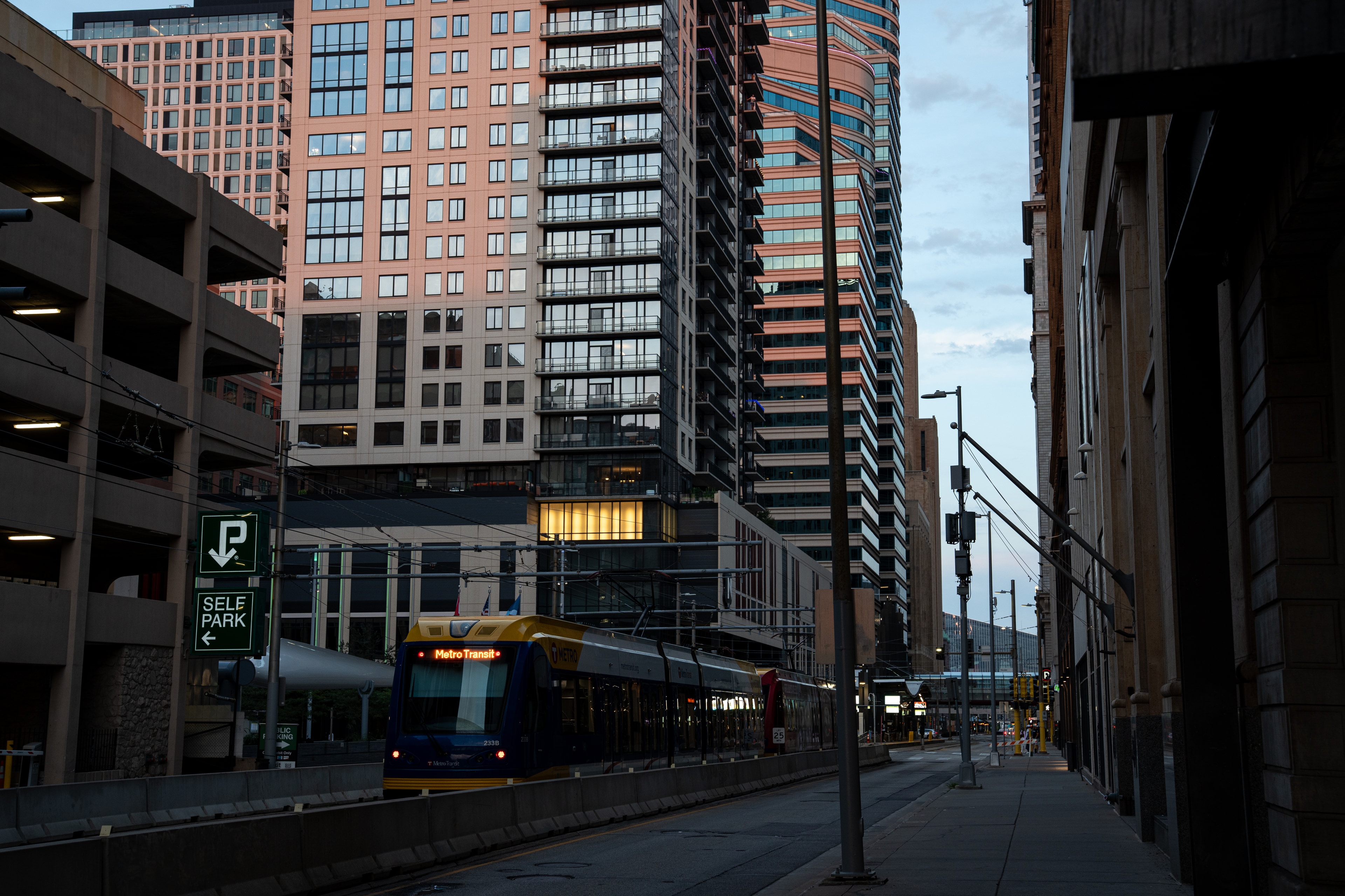 A bus pulling away beneath residential towers at dusk, the street falling into shadow.